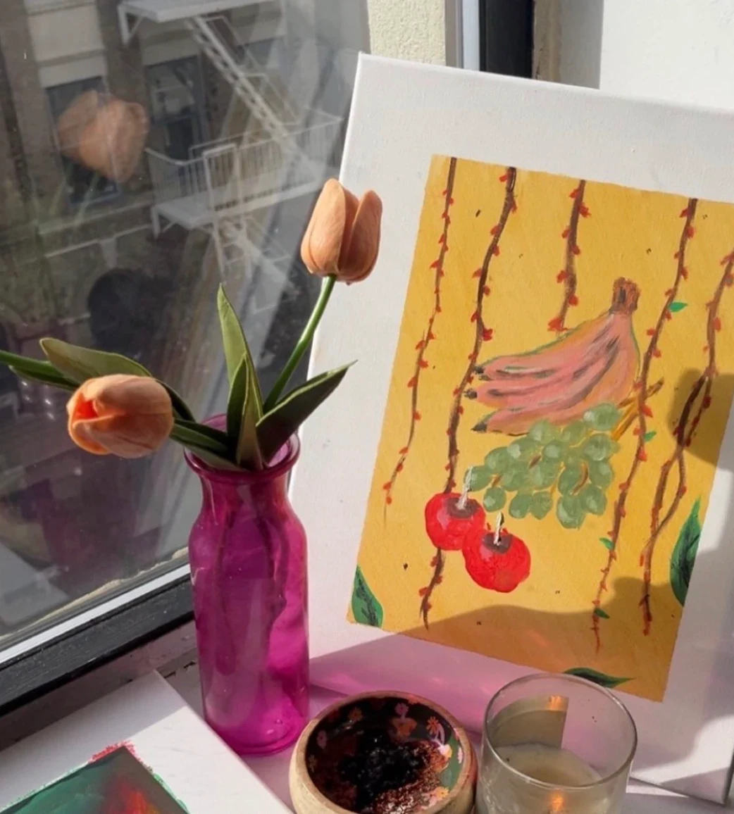 Painting of bananas, green grapes and red cherries hanging from thorny vines on a mustard yellow ground, standing on a windowsill beside a pink vase of tulips.