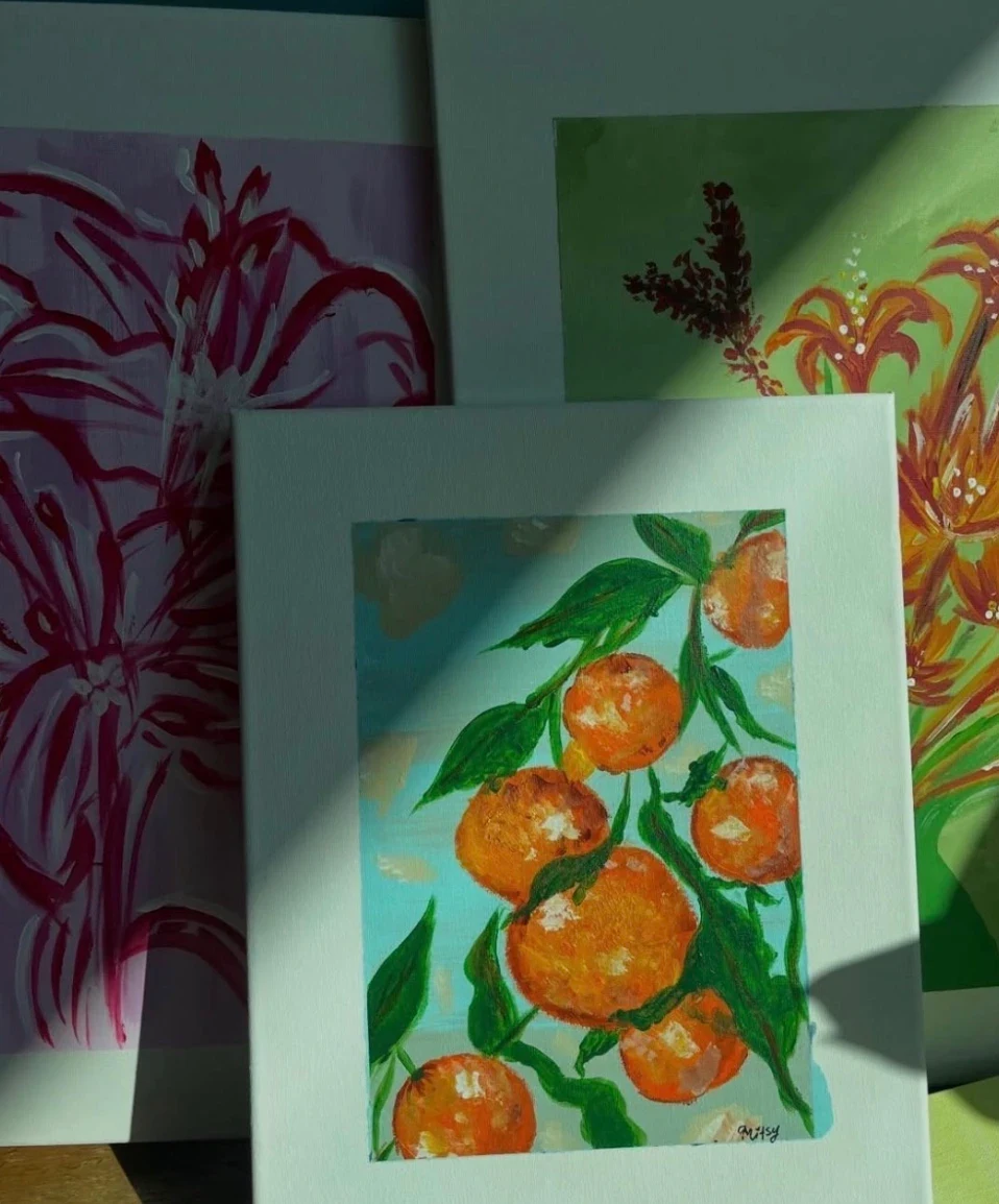 Painting of bright oranges and green leaves against a pale blue sky, signed Mitsy, leaning against two larger flower paintings.