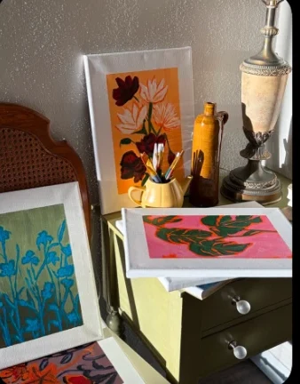 A corner of the studio: paintings of red poppies, pink leaves and blue flowers stacked on and around a small olive green dresser.